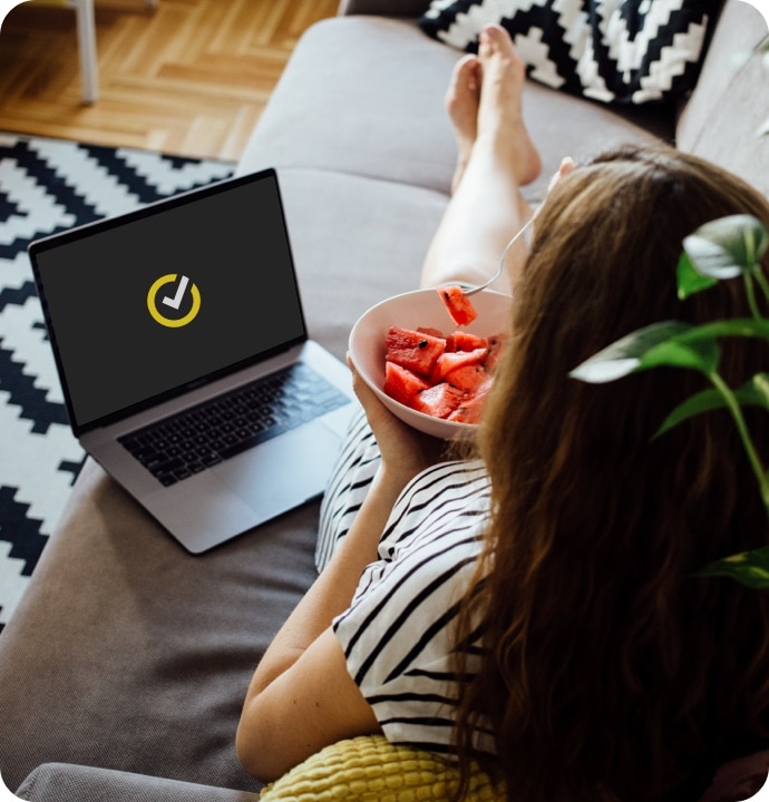 Mujer sobre un sofá con un tazón de fruta y una portátil a su lado con el logotipo de Norton